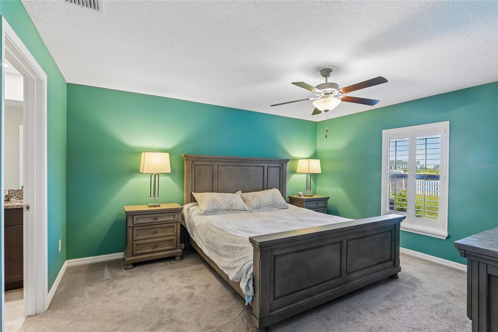 Primary bedroom with carpet flooring, ceiling fan, and plantation shutters on a window with an exterior view. En suite bath access visible through the open doorway; room accommodates a king-size bed with space for additional furniture.