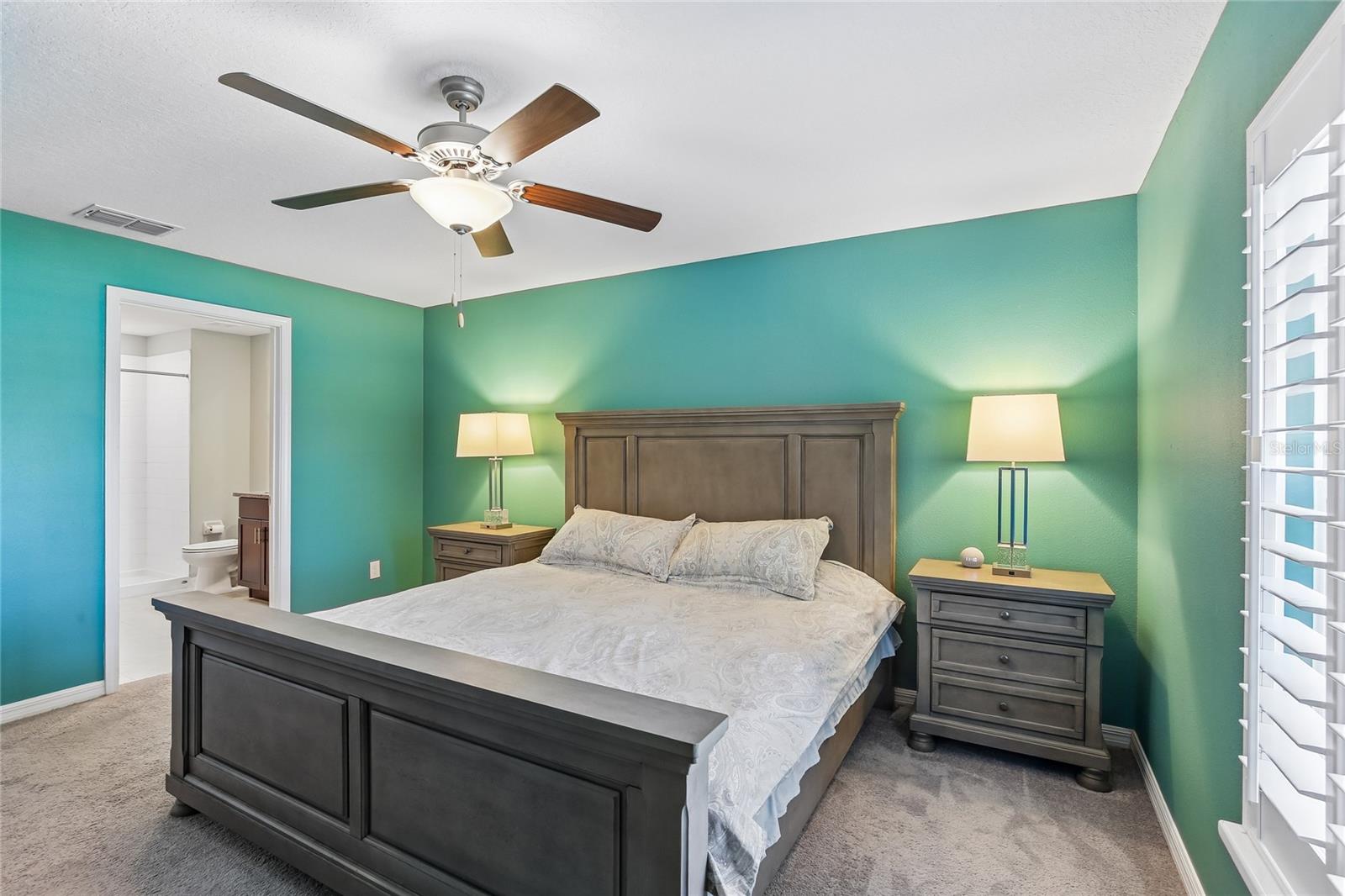 Primary bedroom with carpet flooring, ceiling fan, and plantation shutters. En suite bath entry visible from this angle, with walk-in closet area beyond the doorway.