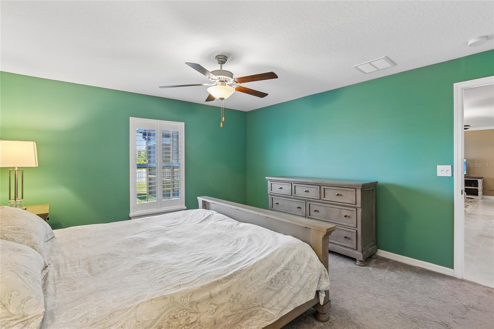 Primary bedroom with carpet flooring, ceiling fan, and a plantation-shuttered window. Doorway connects to the main living area; room size accommodates a king bed and full dresser.