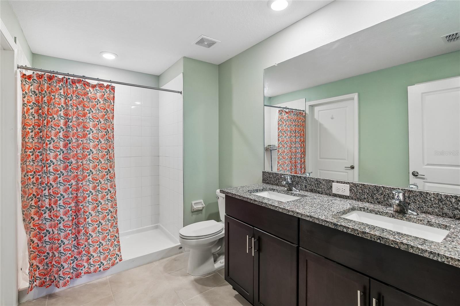 Primary BathPrimary bathroom with a double-sink vanity, espresso cabinetry, granite countertops, full-width mirror, and a walk-in shower with white subway tile surround. Tile flooring and recessed lighting throughout.