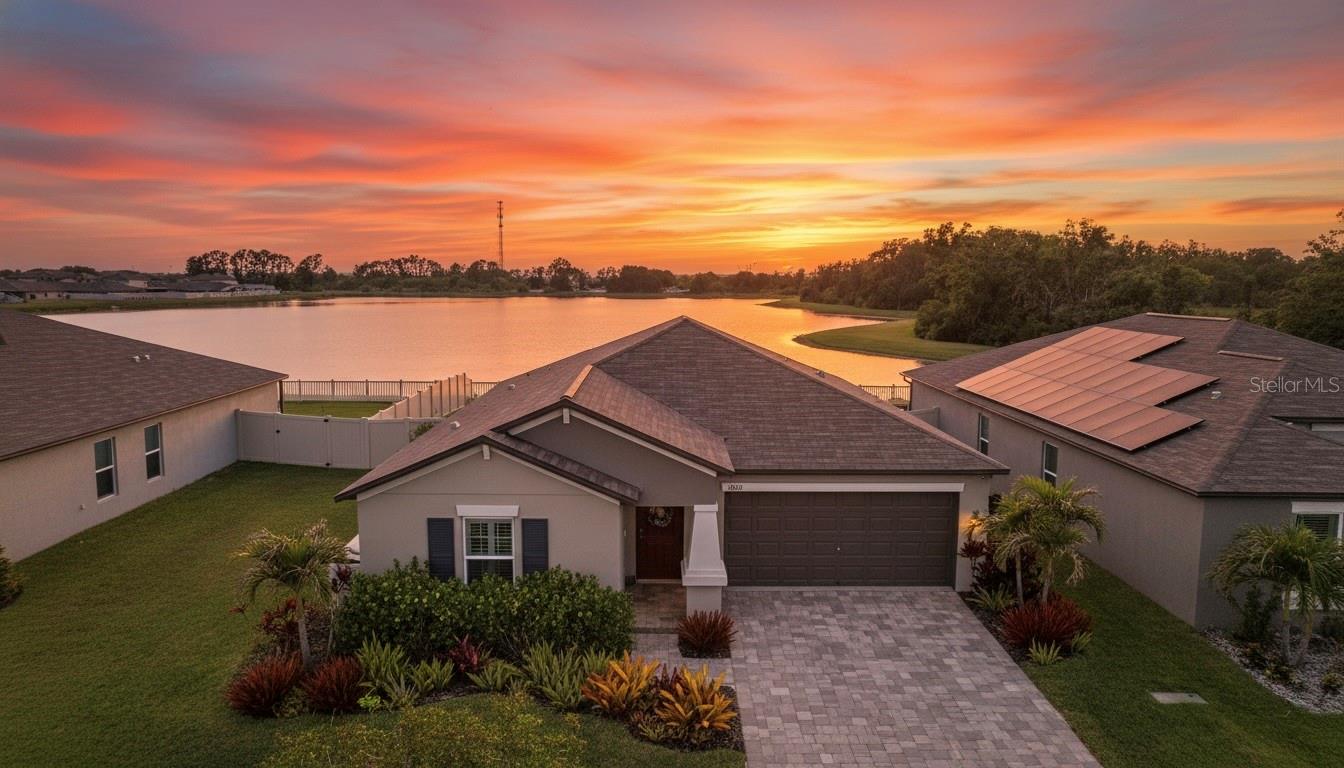 Elevated view of the home's stucco exterior, pavered driveway, and tropical landscaping with the rear pond and mature tree line visible behind the property. Sky digitally enhanced.