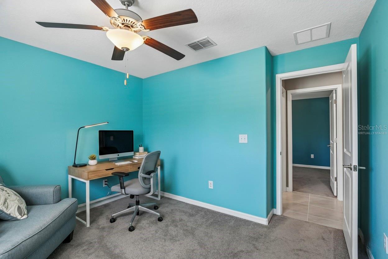 Second bedroom with carpet flooring and ceiling fan, shown virtually staged as a home office. Doorway connects to the hallway; room size accommodates a flex use including bedroom, office, or den.