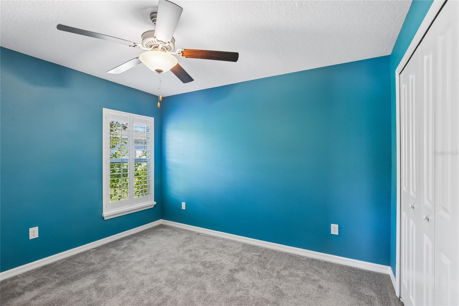 Third bedroom with carpet flooring, ceiling fan, plantation shutters, and a reach-in closet with bifold doors.