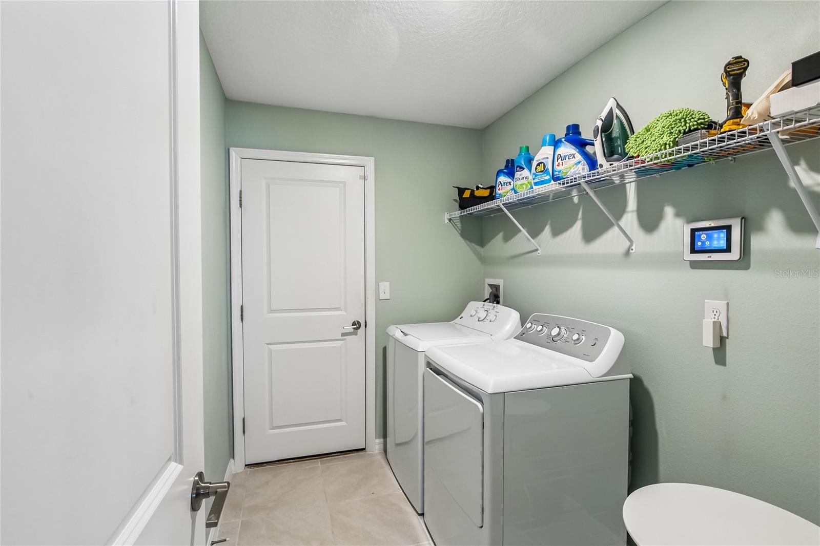 Interior laundry room with tile flooring, full-size washer and dryer, wire shelving above the units, and a door to the garage. Smart home panel visible on the wall.
