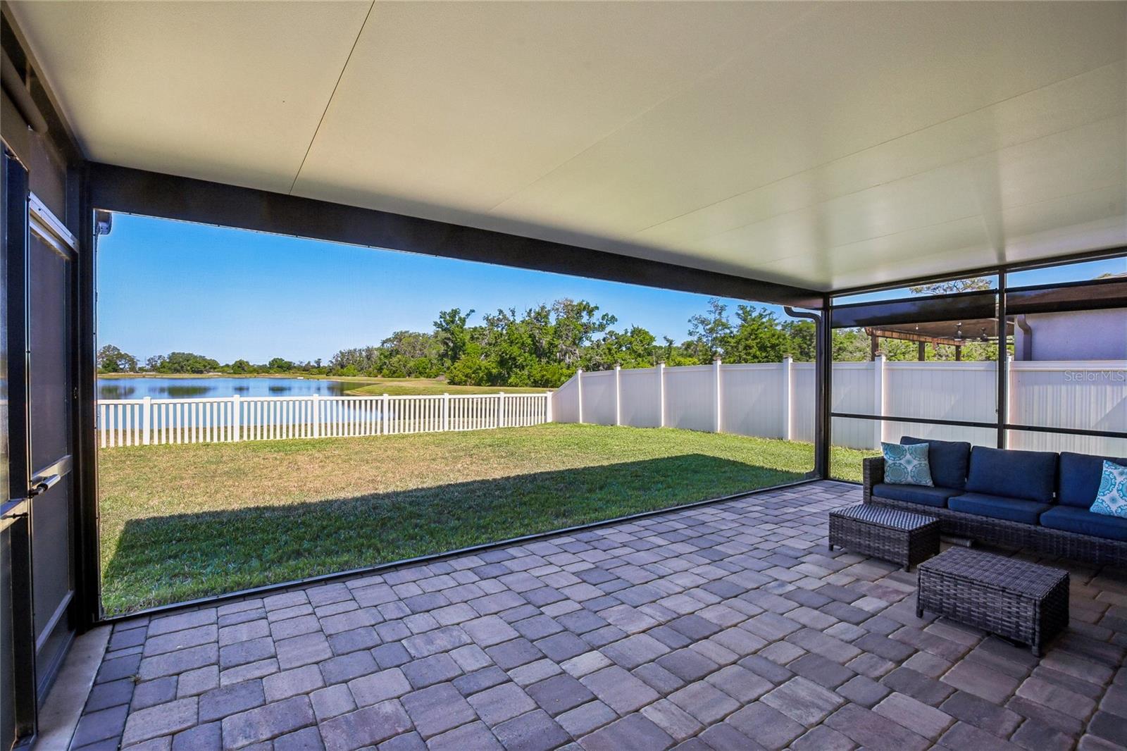 Screened lanai with a pavered floor and open pond view through the screen enclosure. Fenced rear yard with mature tree line visible beyond the water.