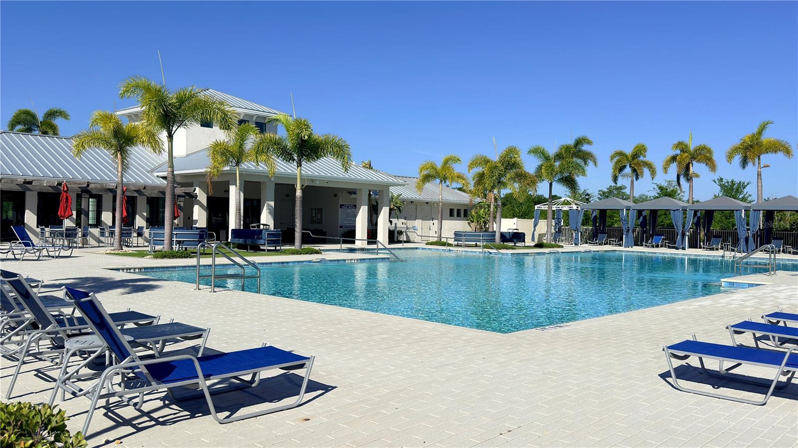 Community resort-style pool with a large pavered deck, lounge chairs, palm trees, covered cabanas, and a spa. Clubhouse with covered outdoor seating area visible in the background.