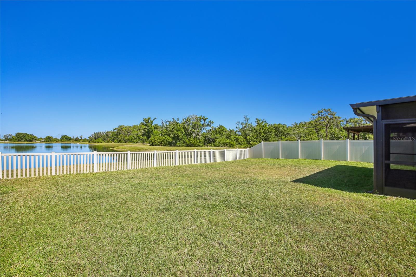 Rear yard with sodded lawn, vinyl fence along the pond line, and a wide open water view. Screened lanai enclosure visible at the right edge of the frame.