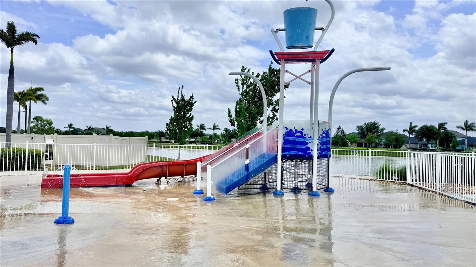Community splash pad with a water slide, dump bucket, and ground-level spray features on a wet-pour surface. Pond view visible in the background beyond the perimeter fence.
