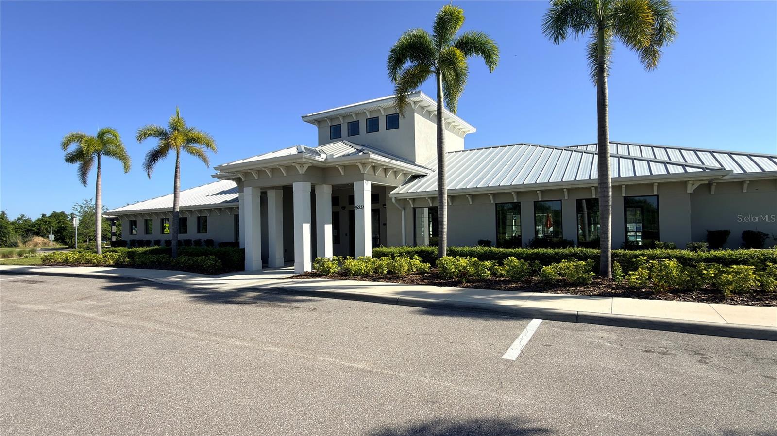 Community clubhouse with a metal roof, covered entry colonnade, floor-to-ceiling windows, and mature royal palm landscaping. Dedicated parking lot on site.