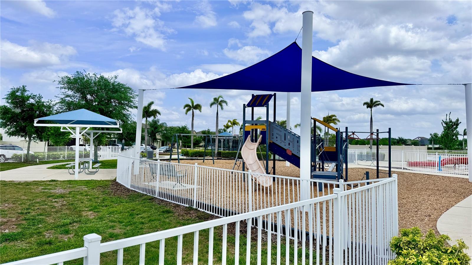 Community playground with a shaded play structure, slide, swing set, and surrounding mulch surface enclosed by a white aluminum fence. Covered picnic area adjacent to the play zone.