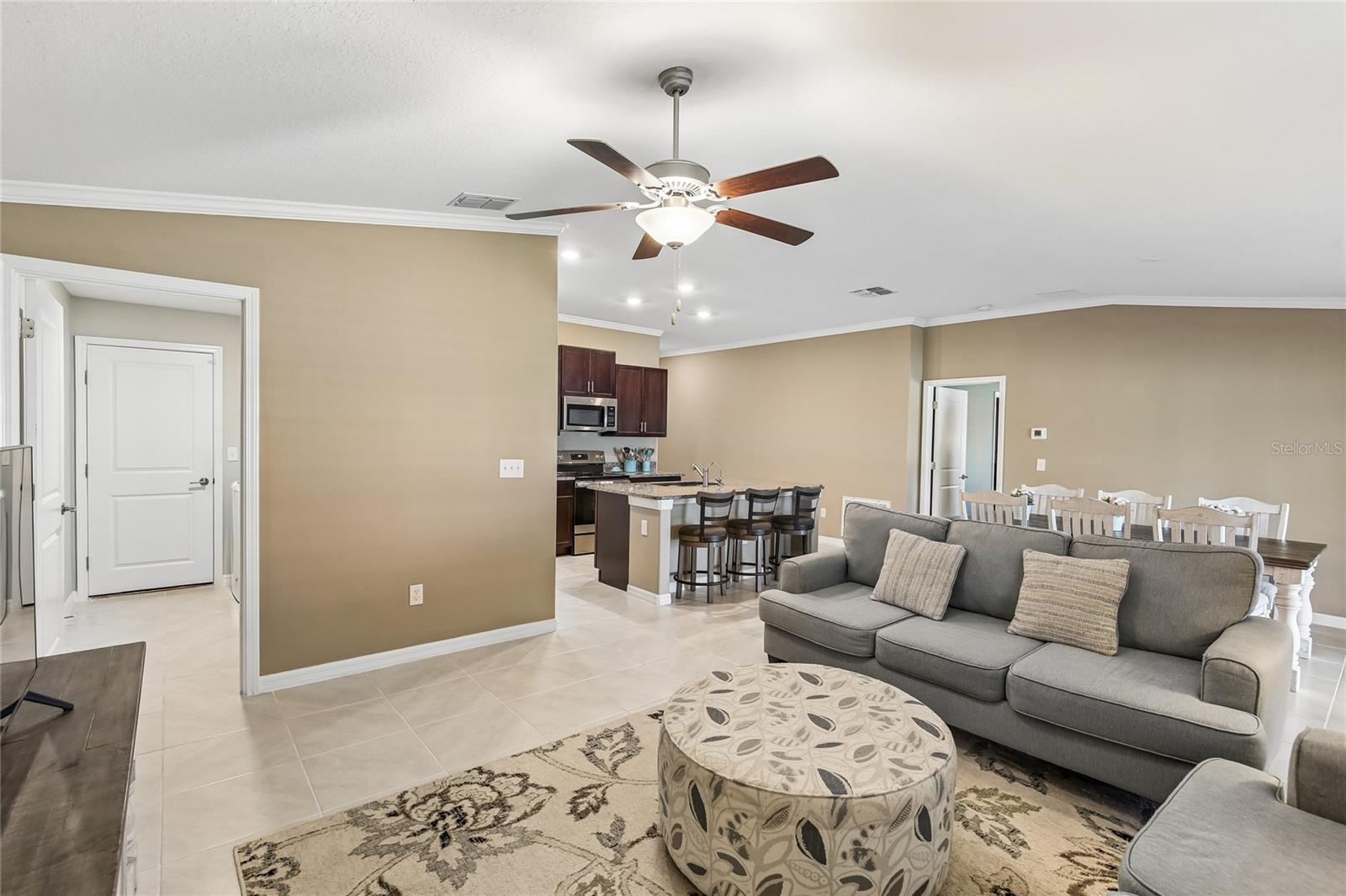 Spacious great room with crown molding, a ceiling fan, and plantation shutters on two windows. Tile flooring and neutral walls flow into an adjacent dining area.