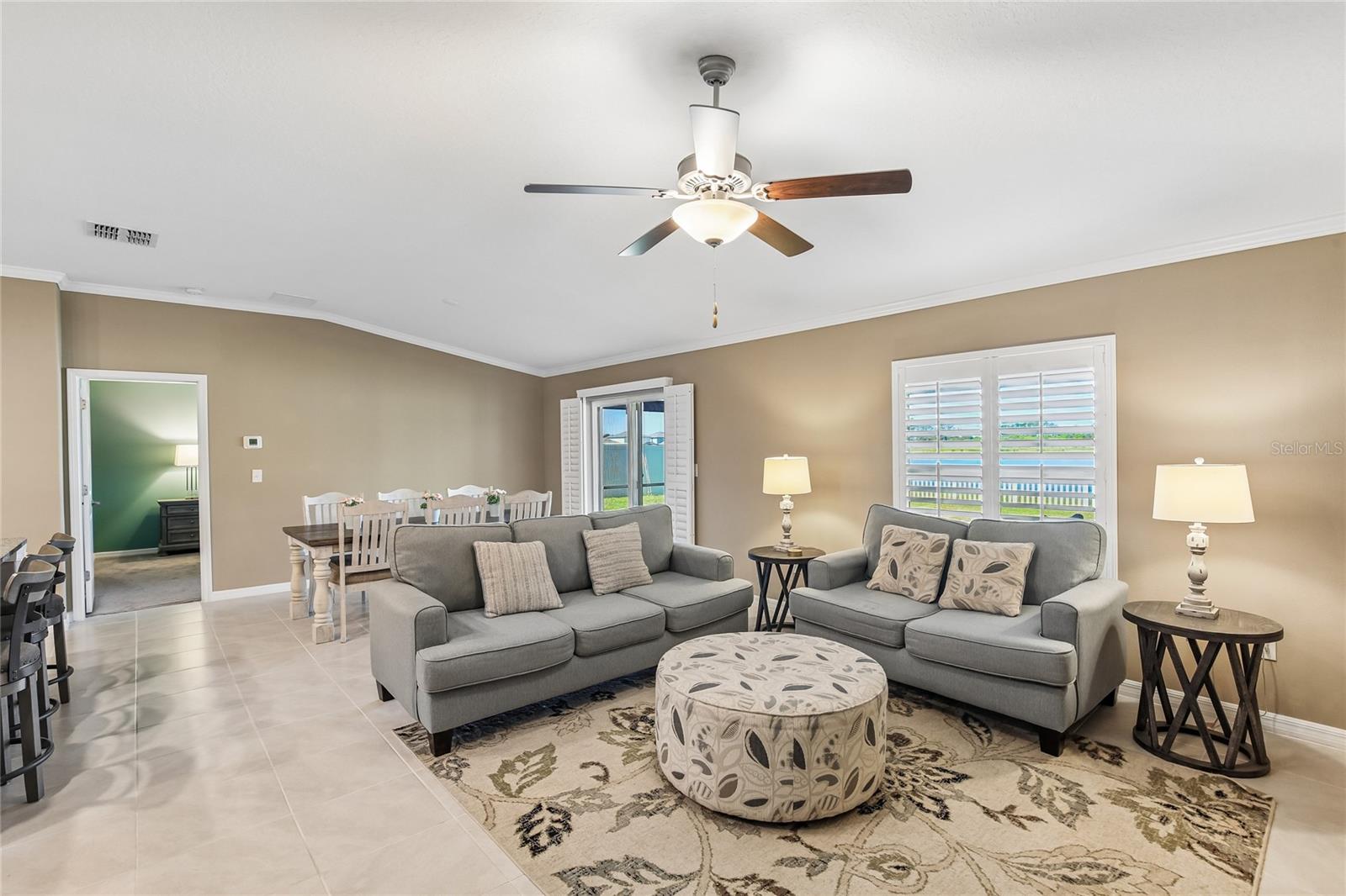 Great room with tile flooring, crown molding, plantation shutters, and a ceiling fan. Room accommodates multiple seating areas and includes a media console along the far wall.