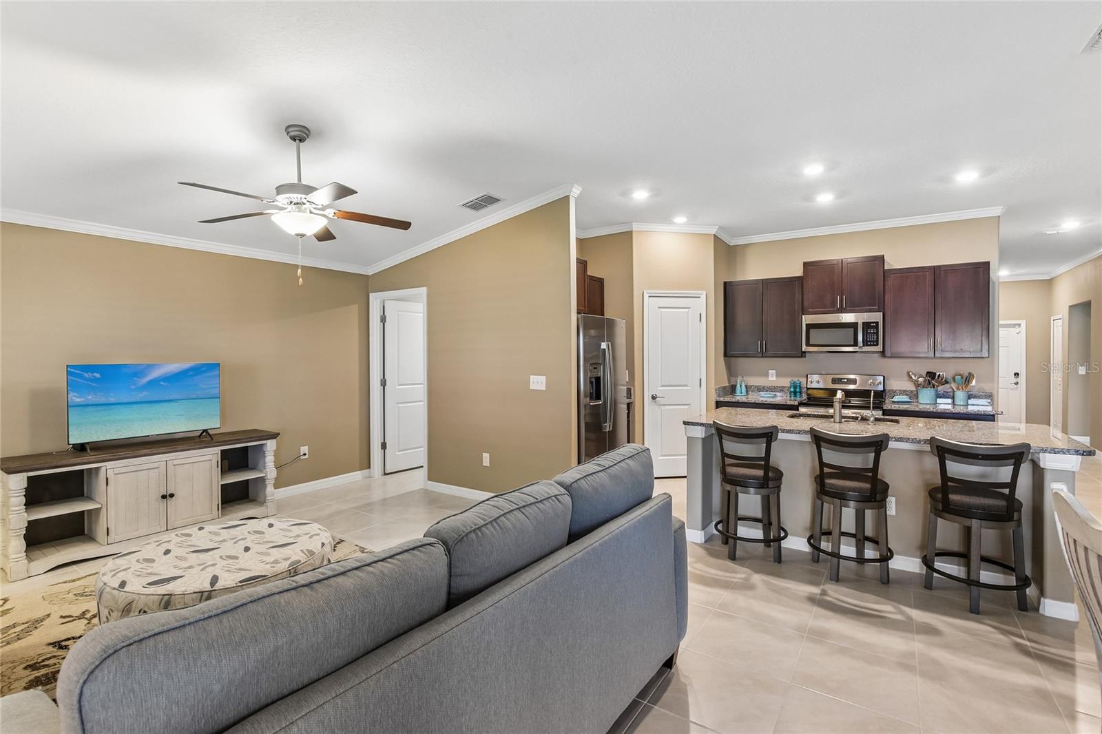 Open-concept living area with tile flooring, crown molding, and a ceiling fan flows directly into the kitchen. Dark espresso cabinetry, granite countertops, a peninsula breakfast bar with barstool seating, and stainless steel appliances are visible from the living space.