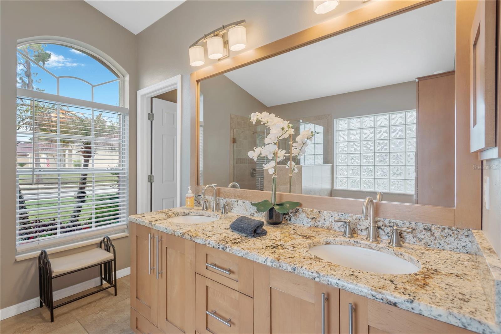 The updated primary en-suite bathroom features dual sinks on a custom built vanity full of storage options and a granite counter. A private water closet is just beyond the vanity.