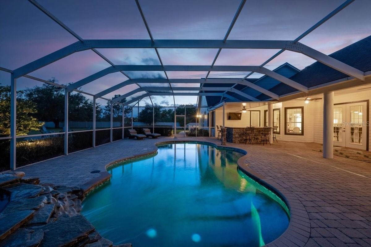Resort-style outdoor oasis featuring a sparkling heated pool and spa, complete with an updated pool pump (2023), heater (2021), and atmospheric pond-style lighting for evening ambiance.