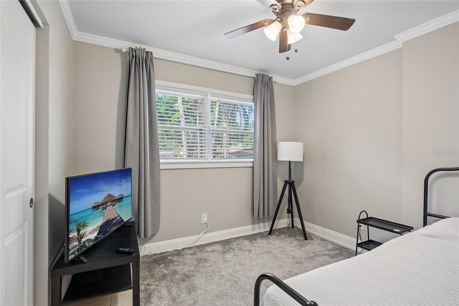 Bedroom Two – Bedroom two offers a comfortable layout with crown molding, a ceiling fan, and a wide window with blinds and curtains. Neutral finishes and soft carpeting create a clean, flexible space.