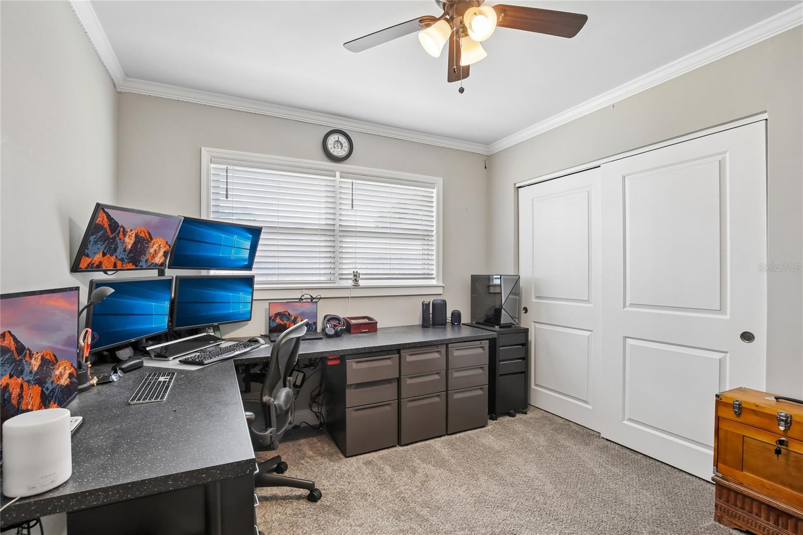 Bedroom Three – Bedroom three is currently arranged as a home office, featuring plush carpeting, crown molding, a ceiling fan, and a wide window with blinds. The room offers flexible use as a bedroom, workspace, or hobby area.