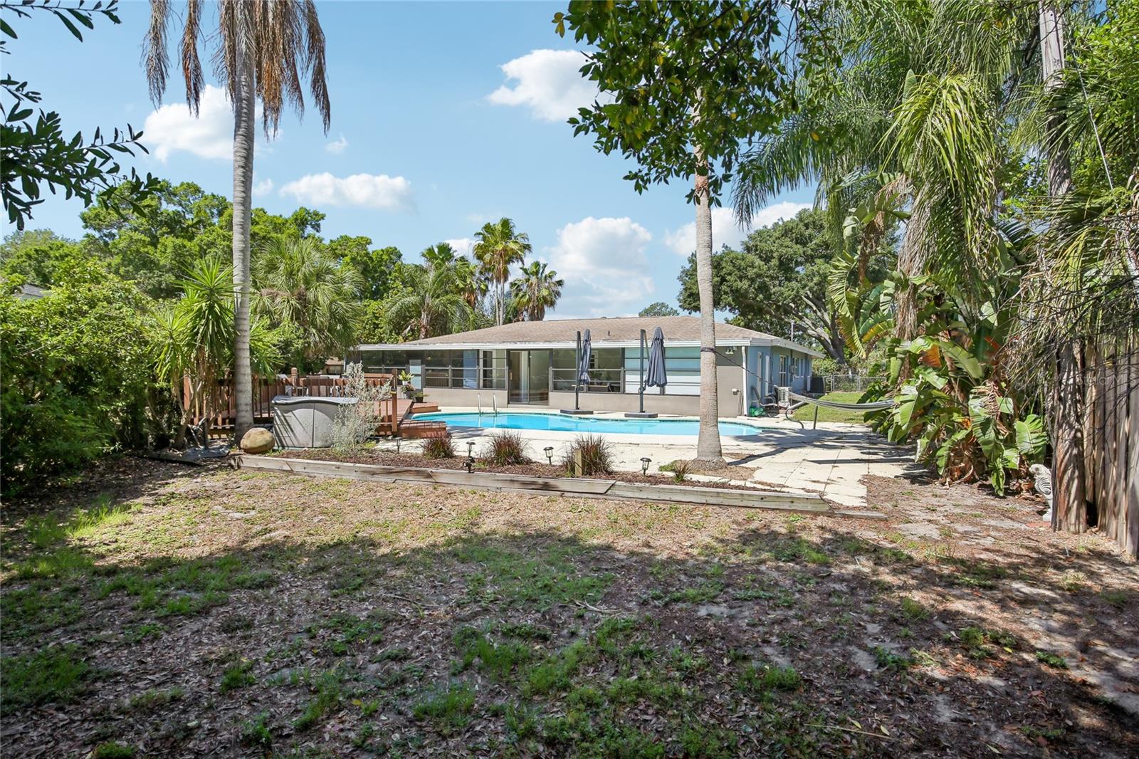 Backyard Overview – This wide backyard view highlights the pool area, raised deck, mature palm trees, and surrounding tropical landscaping. The fenced yard provides a private outdoor setting with room for both relaxing and entertaining.