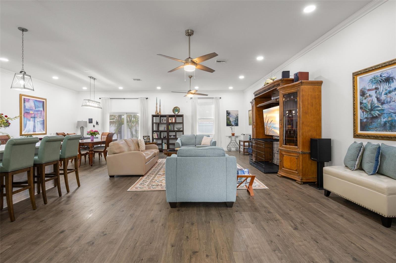 The spacious great room is anchored by wide-plank luxury vinyl flooring, crown molding, recessed lighting, and dual ceiling fans, creating a comfortable gathering space with clean sightlines to the kitchen and dining areas. Sliding glass doors and oversized windows contribute natural light throughout the open interior.