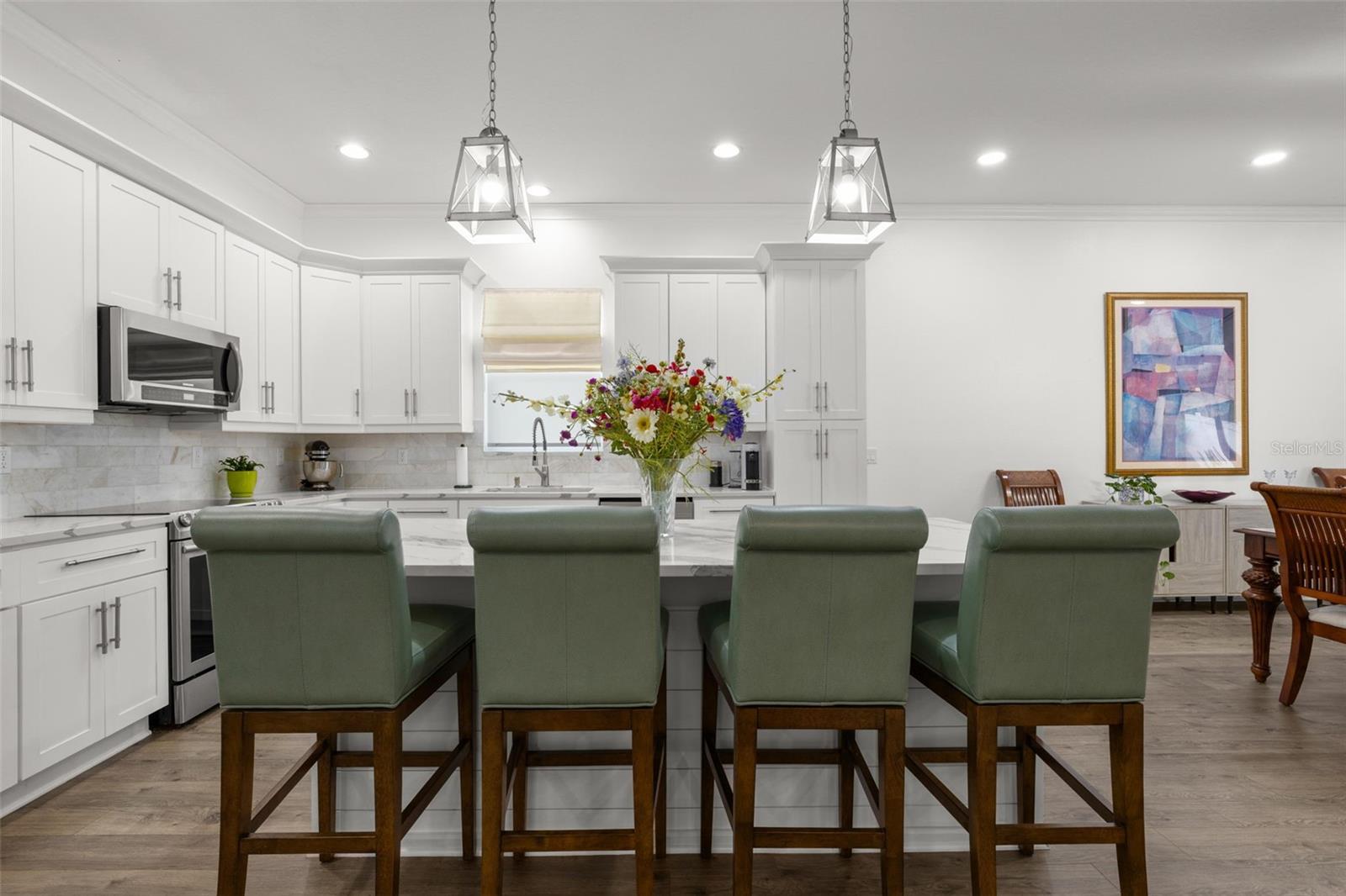 The oversized center island provides extensive prep space, additional storage, integrated microwave, and comfortable seating beneath contemporary pendant lighting. Crisp white shaker cabinetry with crown molding, designer quartz countertops, upgraded stainless appliances, recessed lighting, and an elegant onyx marble backsplash create a clean, sophisticated kitchen design.
