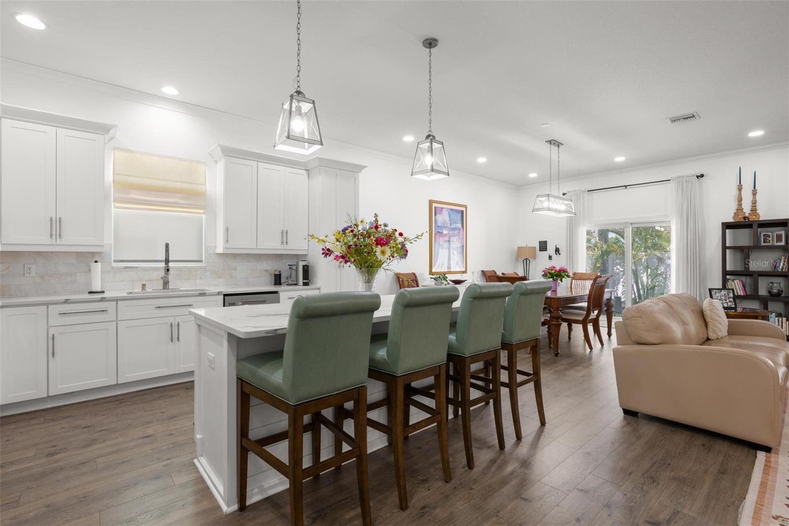 The oversized island offers generous seating and prep space beneath contemporary pendant lighting, while the surrounding open layout connects seamlessly to the dining and living areas. White shaker cabinetry with crown molding, designer quartz countertops, recessed lighting, and wide-plank luxury vinyl flooring continue throughout the bright, cohesive interior.