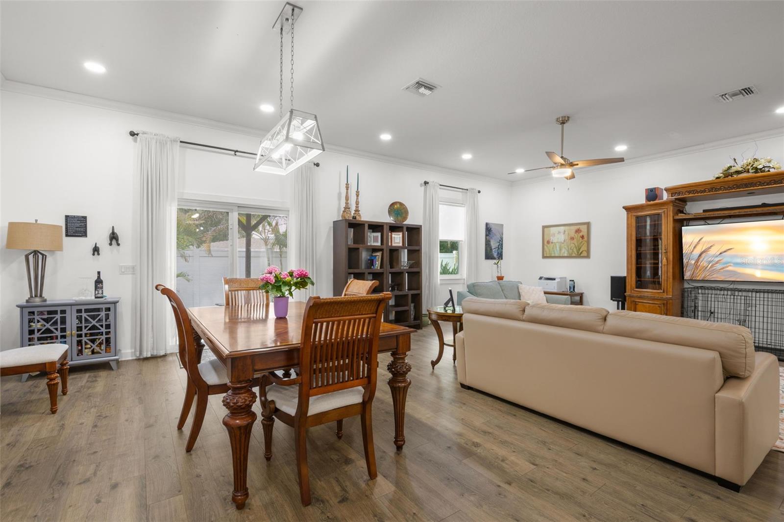 Oversized impact sliding glass doors and large windows fill the dining and living areas with natural light, complementing the home’s high ceilings, crown molding, recessed lighting, and wide-plank luxury vinyl flooring. The open layout offers generous wall space and flexible furniture placement throughout the main gathering areas.