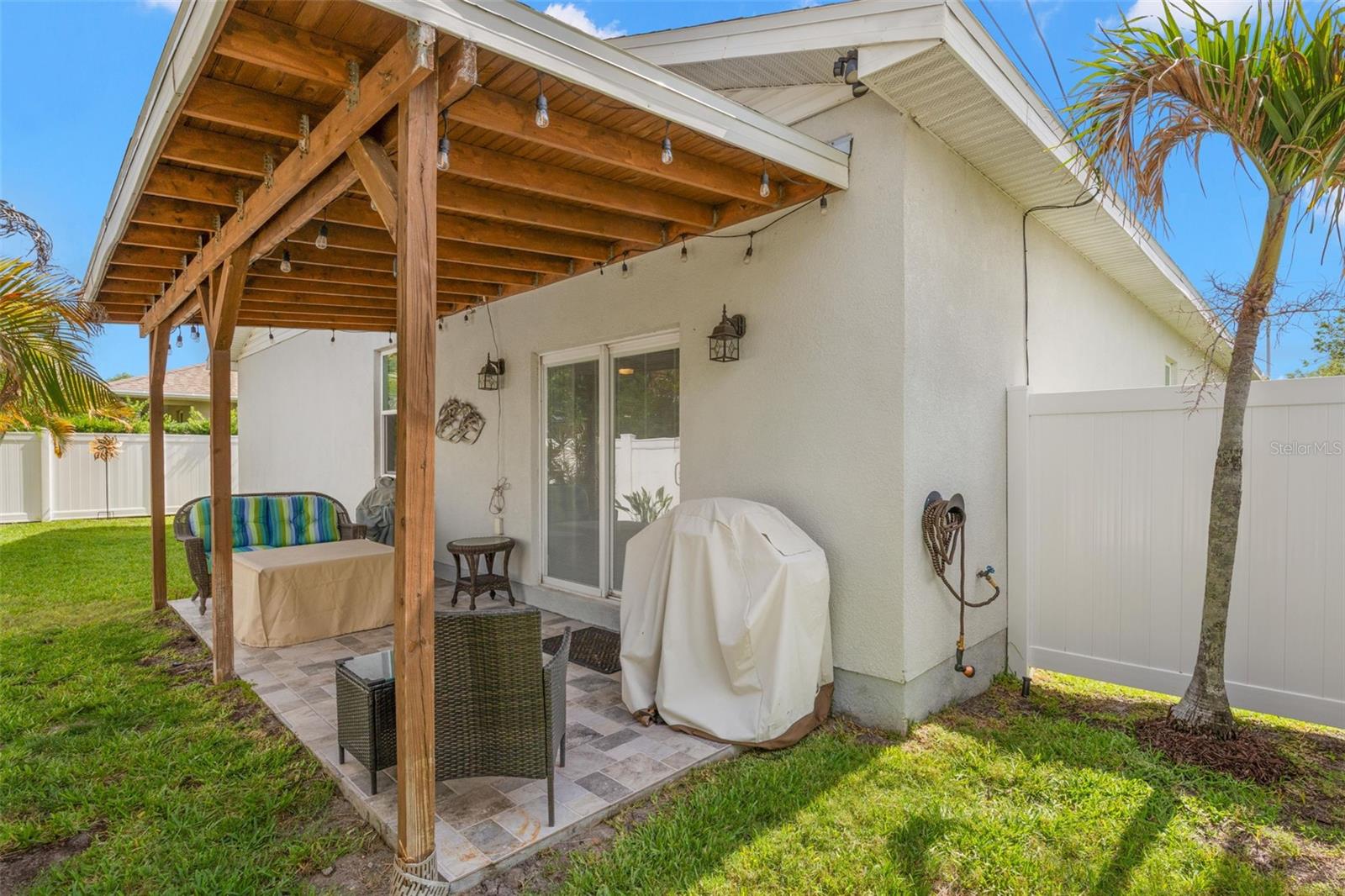The covered rear patio extends the living space outdoors with a warm wood ceiling structure, decorative string lighting, ceramic tile flooring, and room for both dining and lounging. Sliding glass doors provide easy access to the interior, while vinyl fencing creates privacy throughout the backyard.
