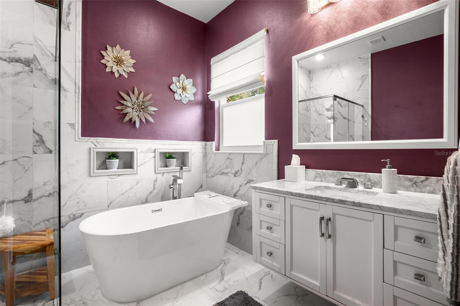 The luxurious primary bath features a freestanding soaking tub, frameless glass walk-in shower with rain shower head, designer quartz countertops, upgraded cabinetry, and dramatic marble-look tile extending throughout the space. Decorative wall niches, upgraded plumbing fixtures, and natural light enhance the spa-like atmosphere.