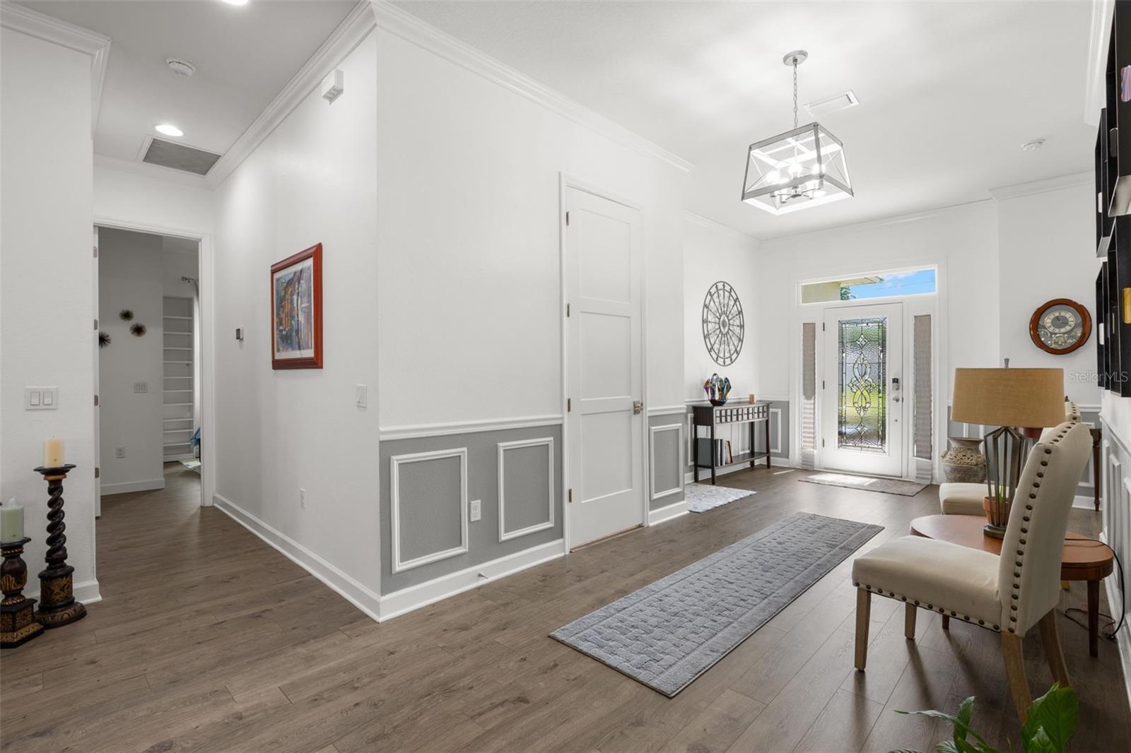 The oversized foyer is framed by extensive crown molding, oversized base trim, wainscoting, chair rail detailing, recessed lighting, and wide-plank luxury vinyl flooring. A statement chandelier, elegant leaded glass entry door with sidelights and transom window, and oversized paneled interior doors contribute to the home’s polished architectural character.