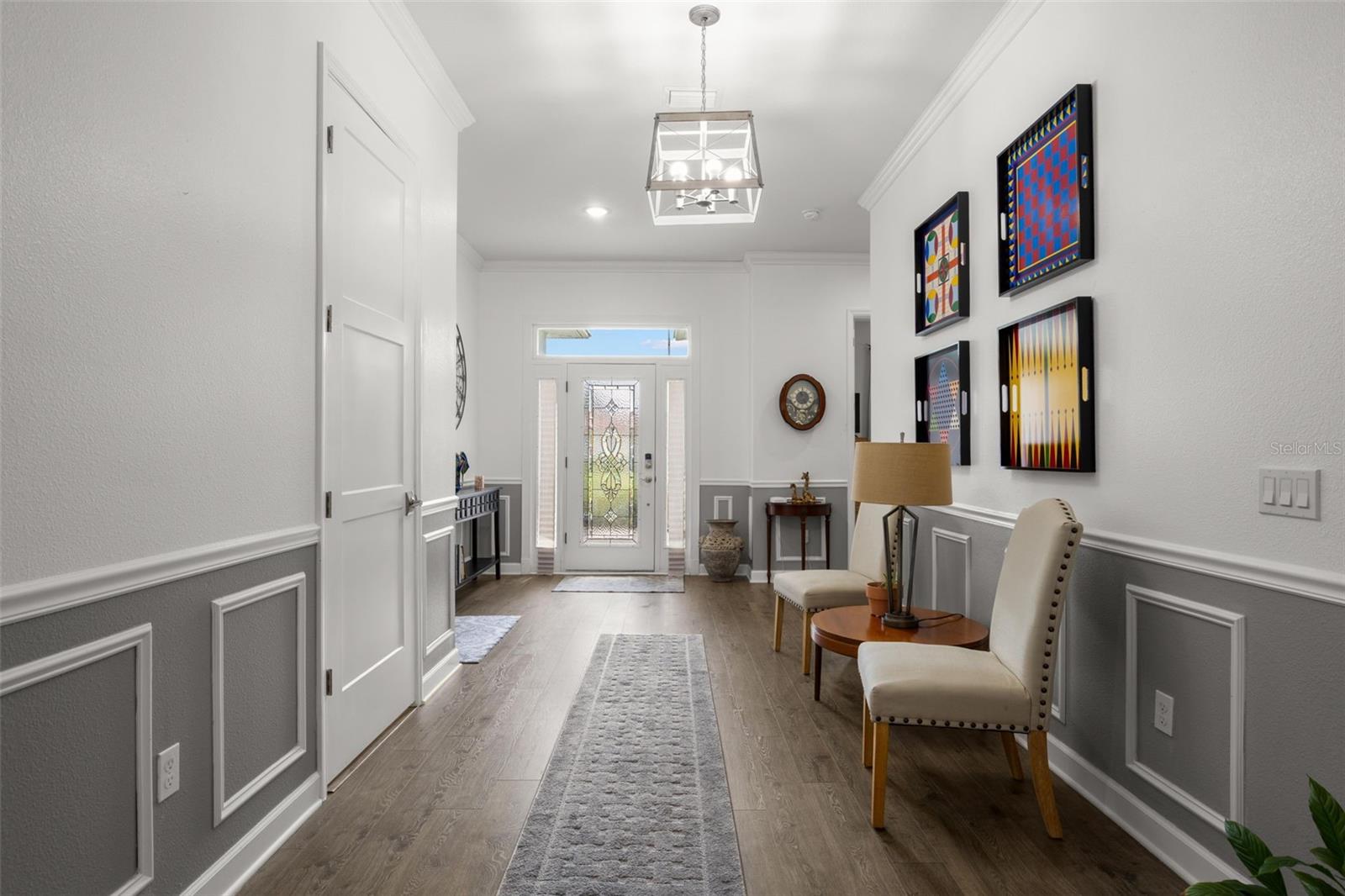 The impressive foyer showcases extensive crown molding, oversized base trim, wainscoting, chair rail detailing, recessed lighting, a statement chandelier, and wide-plank luxury vinyl flooring. A leaded glass entry door with sidelights and transom window adds natural light and architectural character to the welcoming entryway.