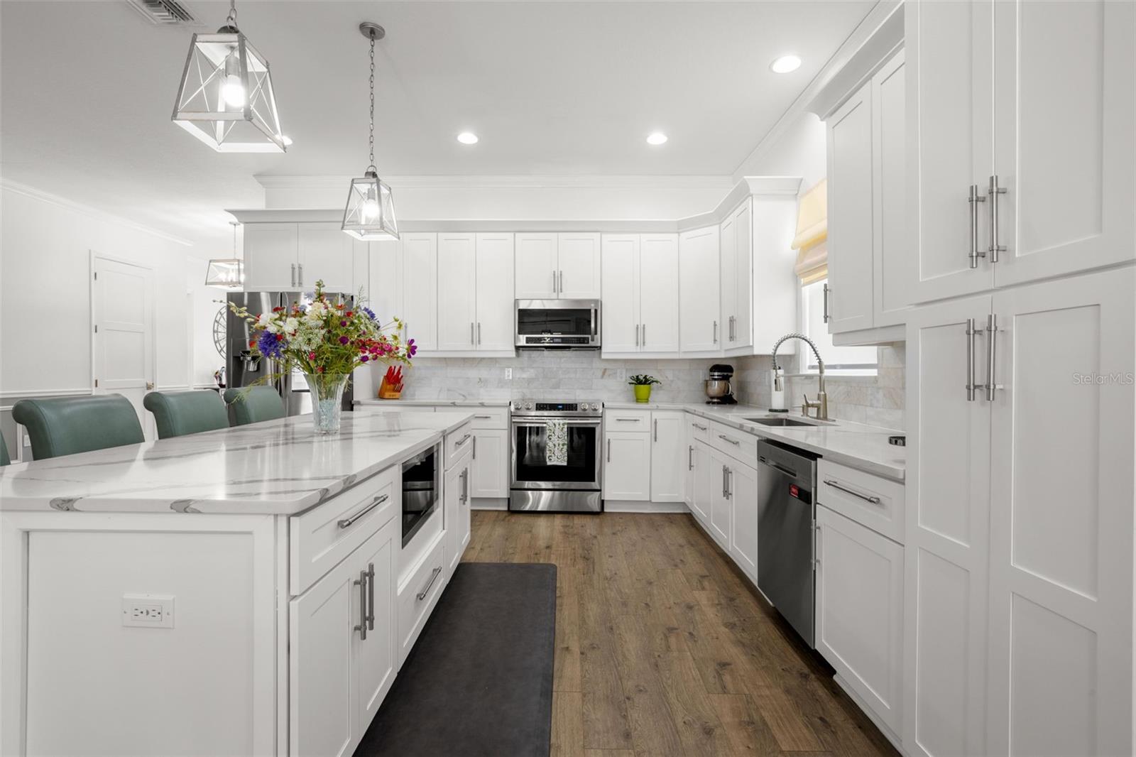 The chef-inspired kitchen combines clean architectural lines with high-end finishes, including white shaker cabinetry with crown molding, designer quartz countertops, an oversized center island with additional storage and seating, upgraded stainless appliances, and an undermount farmhouse sink. An elegant onyx marble backsplash, pendant lighting, recessed lighting, and extensive cabinetry complete the beautifully designed space.