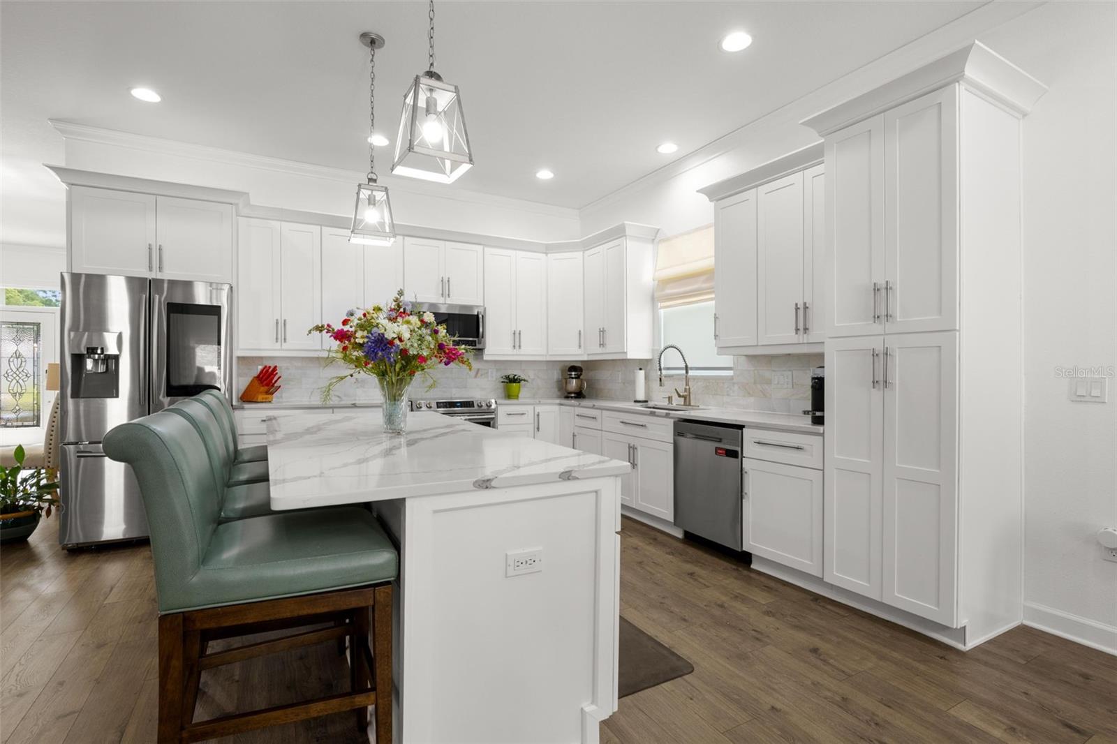 The designer kitchen features white shaker cabinetry with oversized crown molding, designer quartz countertops, a dramatic oversized center island with seating, upgraded stainless appliances, and an undermount farmhouse sink. Pendant lighting, recessed lighting, an onyx marble backsplash, and abundant cabinetry elevate both the functionality and visual appeal of the space.