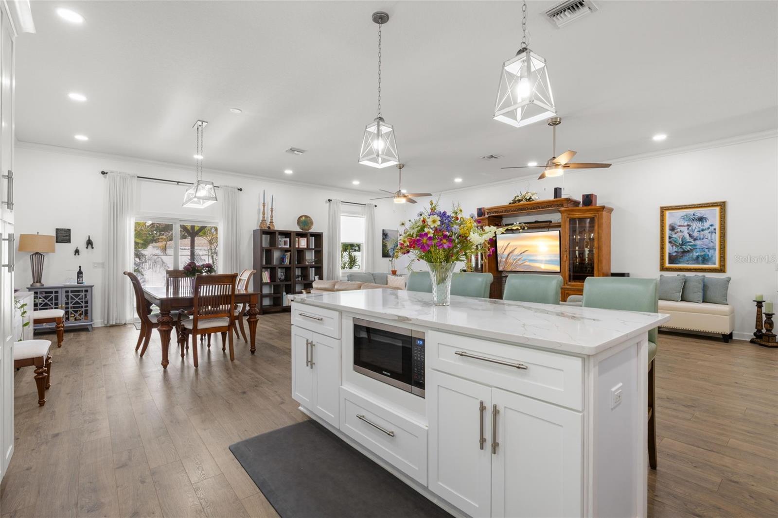 The oversized center island serves as a striking focal point within the open-concept living space, offering designer quartz countertops, additional storage, integrated 2nd microwave, pendant lighting, and generous seating. Open sightlines connect the kitchen, dining area, and great room beneath high ceilings with recessed lighting and dual contemporary ceiling fans.