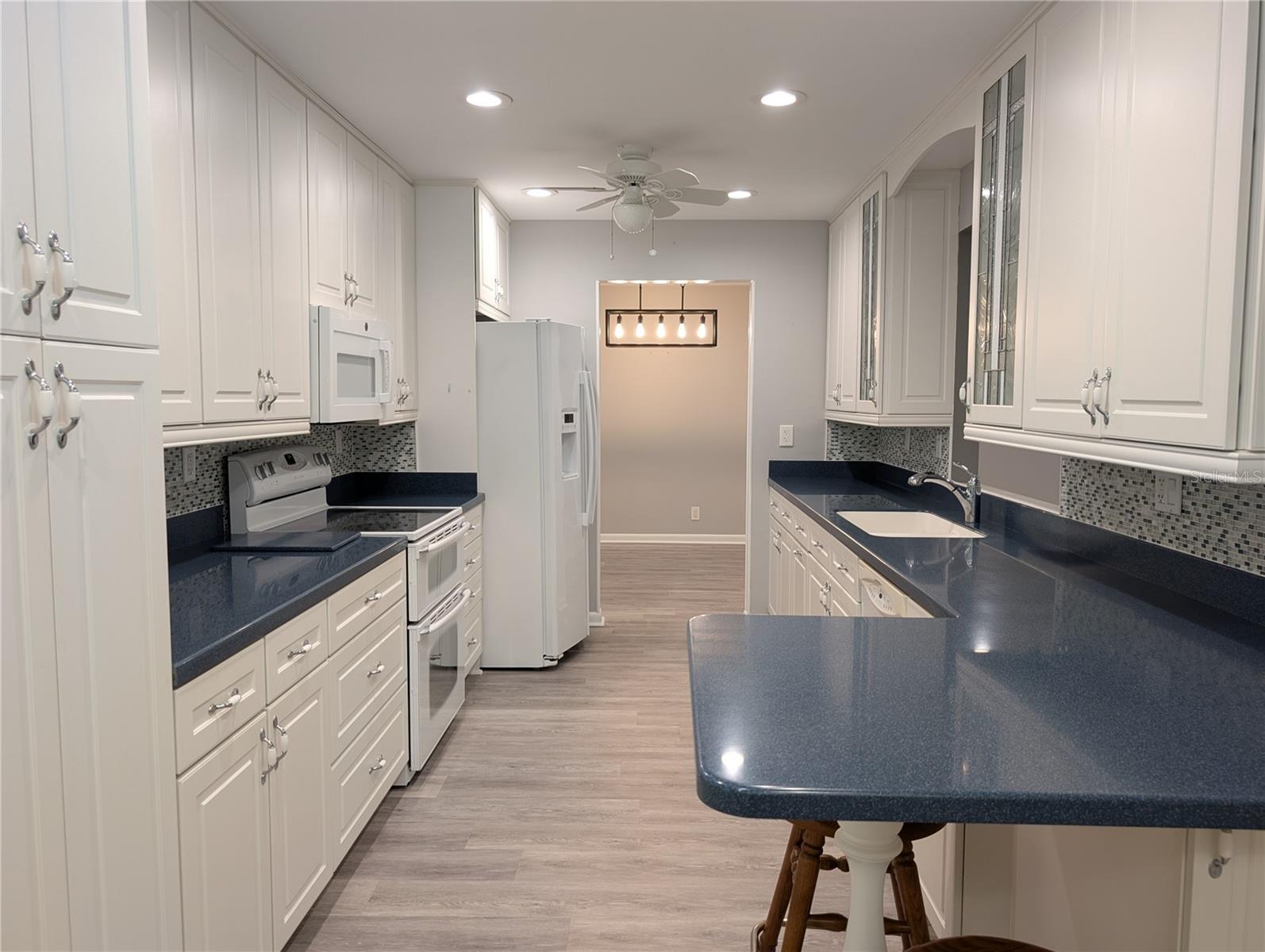 View of kitchen into dining room. Family room to right. Stunning kitchen features elongated counterspace, luxury vinyl flooring (2023), white 42" Kraftmaid cabinets with cobalt blue solid surface countertops. Tall ceiling with recessed lighting and two ceiling fans.