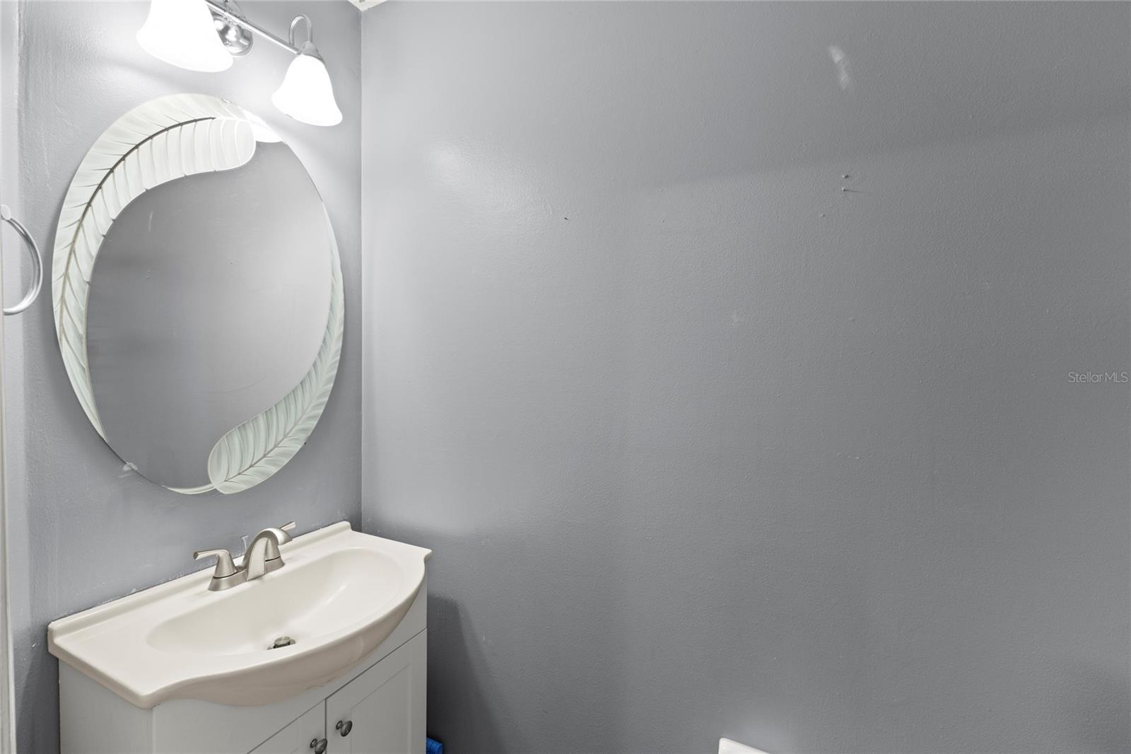 Main floor powder room featuring a sculptural curved mirror with etched-glass detailing, dual-light vanity fixture, brushed-nickel faucet, compact vanity with cabinet storage, and neutral paint tones. Conveniently located on the first level for guests.
