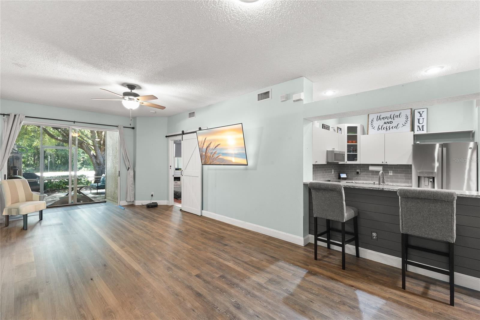 Open living and dining area with wood-look laminate flooring, soft neutral tones, ceiling fan with light kit, recessed lighting, and sliding glass doors leading to the screened rear porch. The layout offers flexibility for multiple furniture arrangements, while the elevated breakfast bar creates a visual connection to the kitchen.