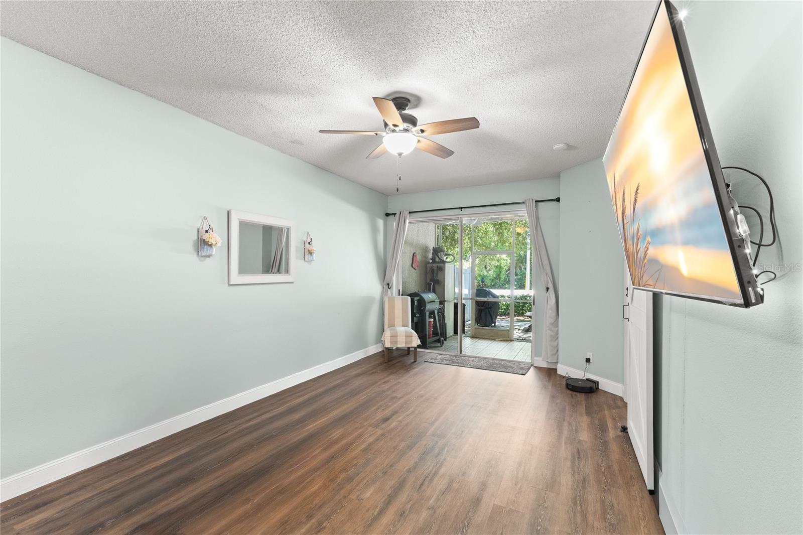 Living area with wood-look laminate flooring, soft neutral tones, ceiling fan with light kit, and sliding glass doors opening to the screened rear porch. The elongated layout provides flexibility for seating arrangements while drawing natural light into the main living space.