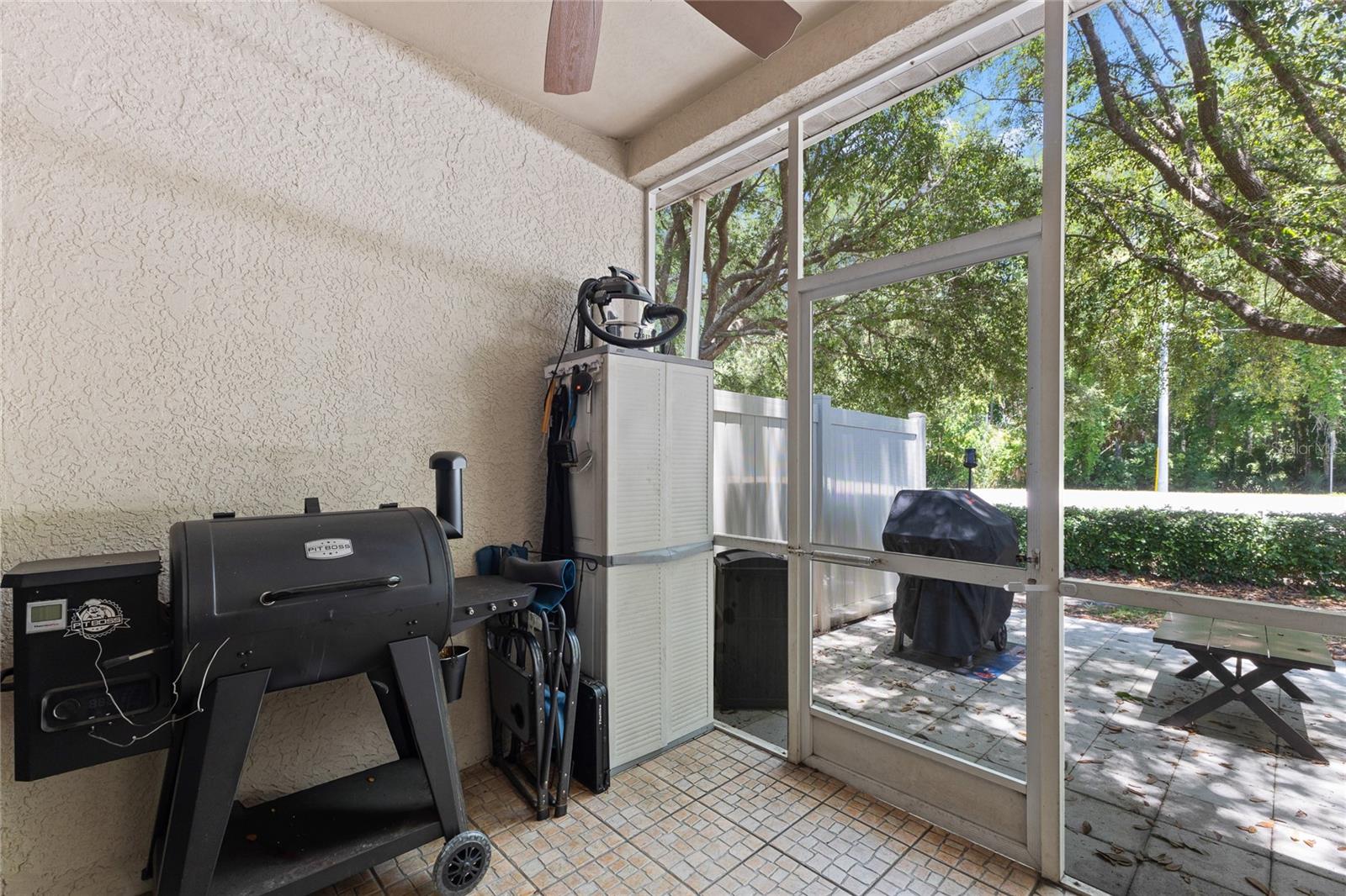 Screened rear porch with tile flooring, ceiling fan, and direct access to the outdoor patio area. Surrounded by mature trees and greenery, the space offers shaded outdoor seating and room for grilling or casual dining.