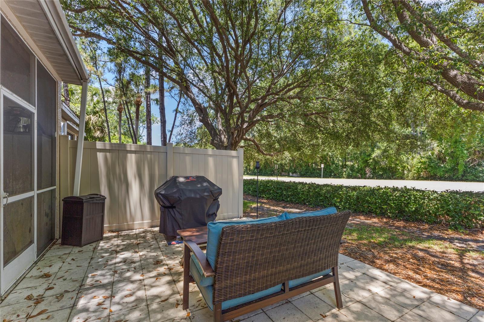 Outdoor patio area adjacent to the screened porch with paver decking, privacy fencing, and mature shade trees creating a natural backdrop. The space offers room for grilling, outdoor seating, and relaxed outdoor living.