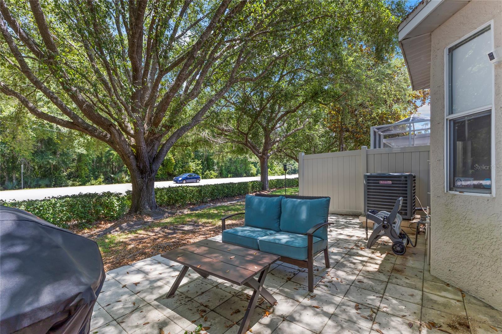 Paver patio bordered by mature shade trees and privacy fencing, creating a shaded outdoor setting with room for grilling, seating, and casual outdoor gatherings. The surrounding greenery adds a natural backdrop and additional privacy.