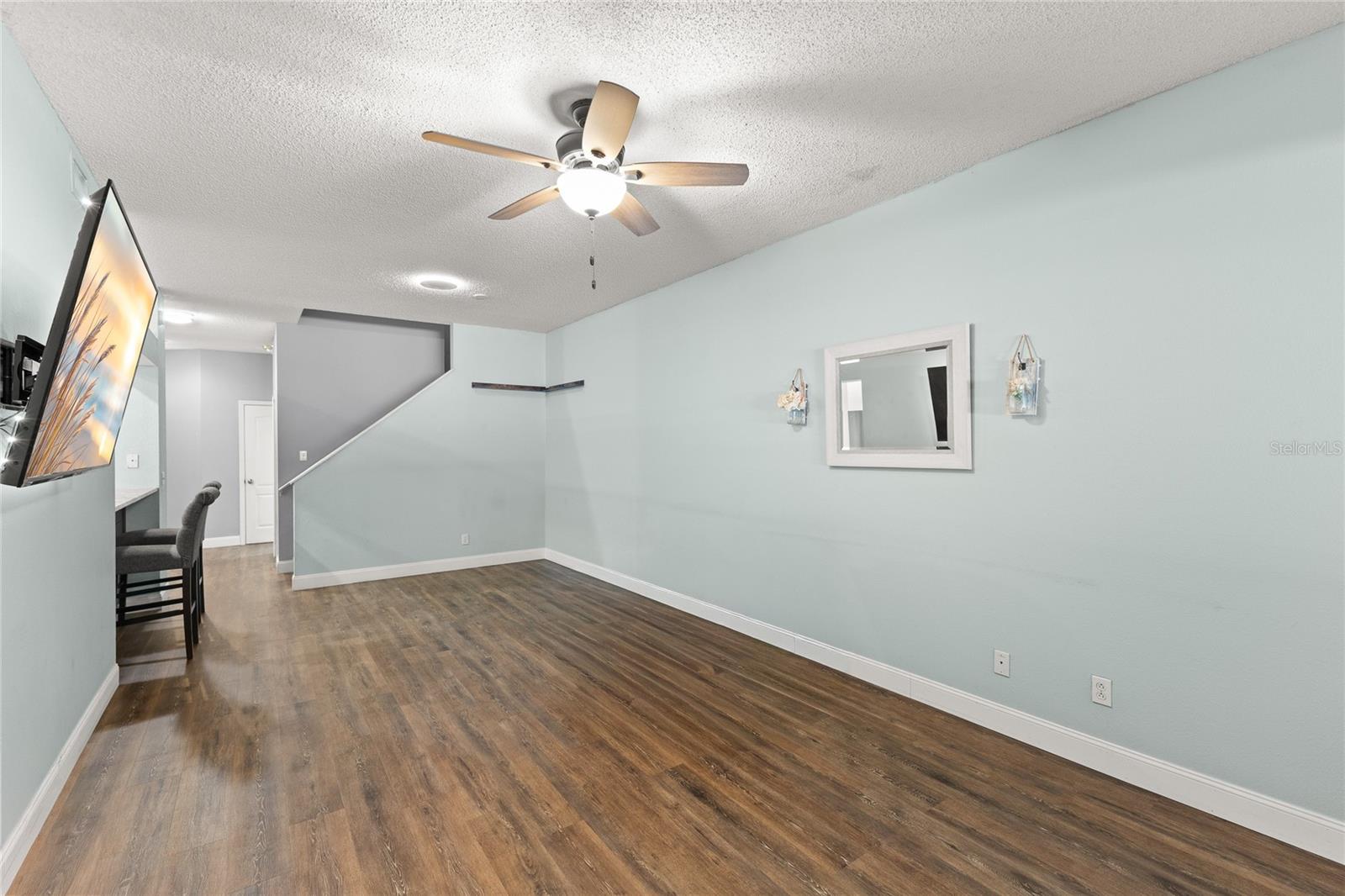 Open living and dining area with wood-look laminate flooring, soft neutral tones, recessed lighting, and a ceiling fan with light kit. The elongated layout provides flexibility for furniture placement and creates clear sightlines toward the kitchen and staircase.