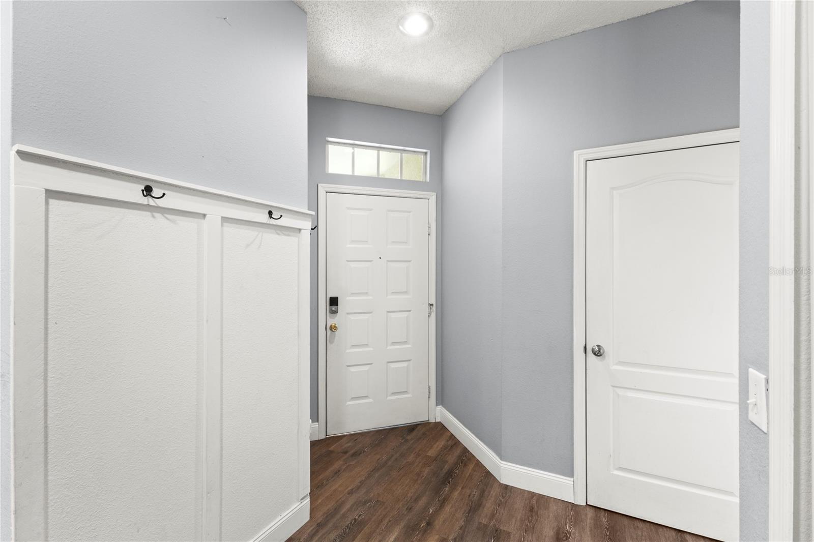 Foyer with soaring ceilings, transom window above the entry door, neutral interior paint, decorative wall trim with coat hooks, and wood-look laminate flooring extending into the main living areas.