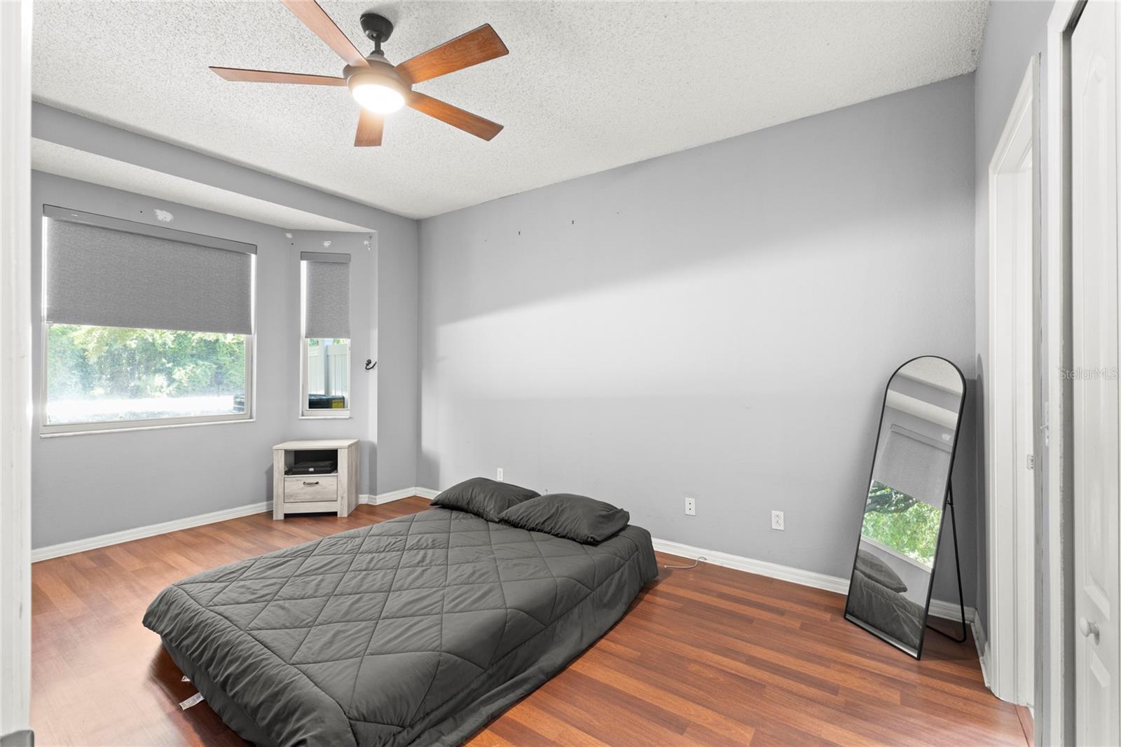 First-floor primary bedroom with warm wood-look flooring, neutral paint tones, ceiling fan with light kit, and a window alcove bringing in natural light and views of the surrounding greenery. The spacious layout offers flexibility for larger furniture pieces and a sitting area or workspace.