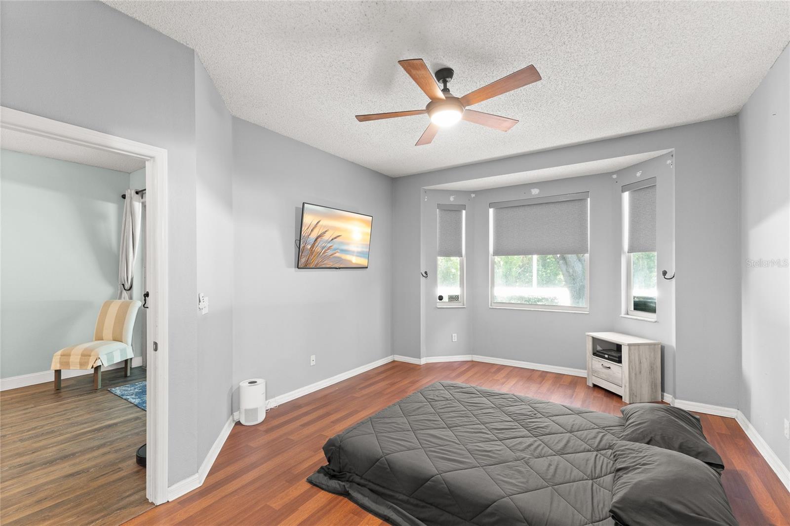 First-floor primary bedroom with warm wood-look flooring, neutral paint tones, ceiling fan with light kit, and a bay-style window alcove bringing in natural light and views of the surrounding greenery. Spacious layout offers flexibility for larger furniture pieces, a sitting area, or workspace.