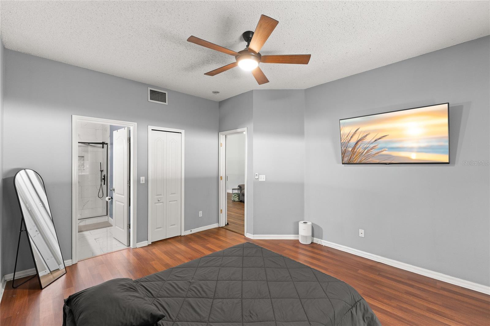 First-floor primary bedroom with warm wood-look flooring, neutral paint tones, and a ceiling fan with light kit. Spacious layout includes a walk-in closet and direct access to the ensuite bath.