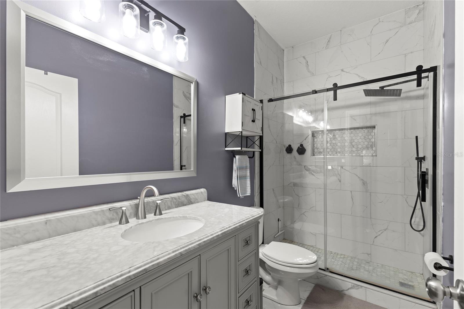 Primary ensuite bath with tile flooring, oversized walk-in shower, marble-look wall tile, frameless sliding glass enclosure with matte black hardware, recessed shower niche with mosaic tile accent, and a rainfall shower head with handheld attachment. The space also features a furniture-style vanity with a marble-look countertop, brushed-nickel fixtures, and contemporary vanity lighting.