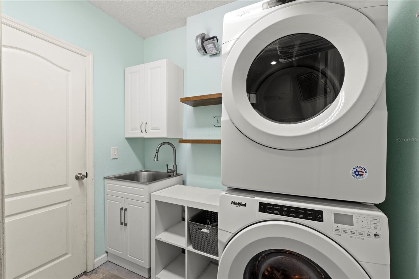 First-floor laundry room with full-size stacked washer and dryer, utility sink, upper cabinet storage, open shelving, and folding/work surface. Functional layout offers additional organization and everyday convenience near the garage entry.