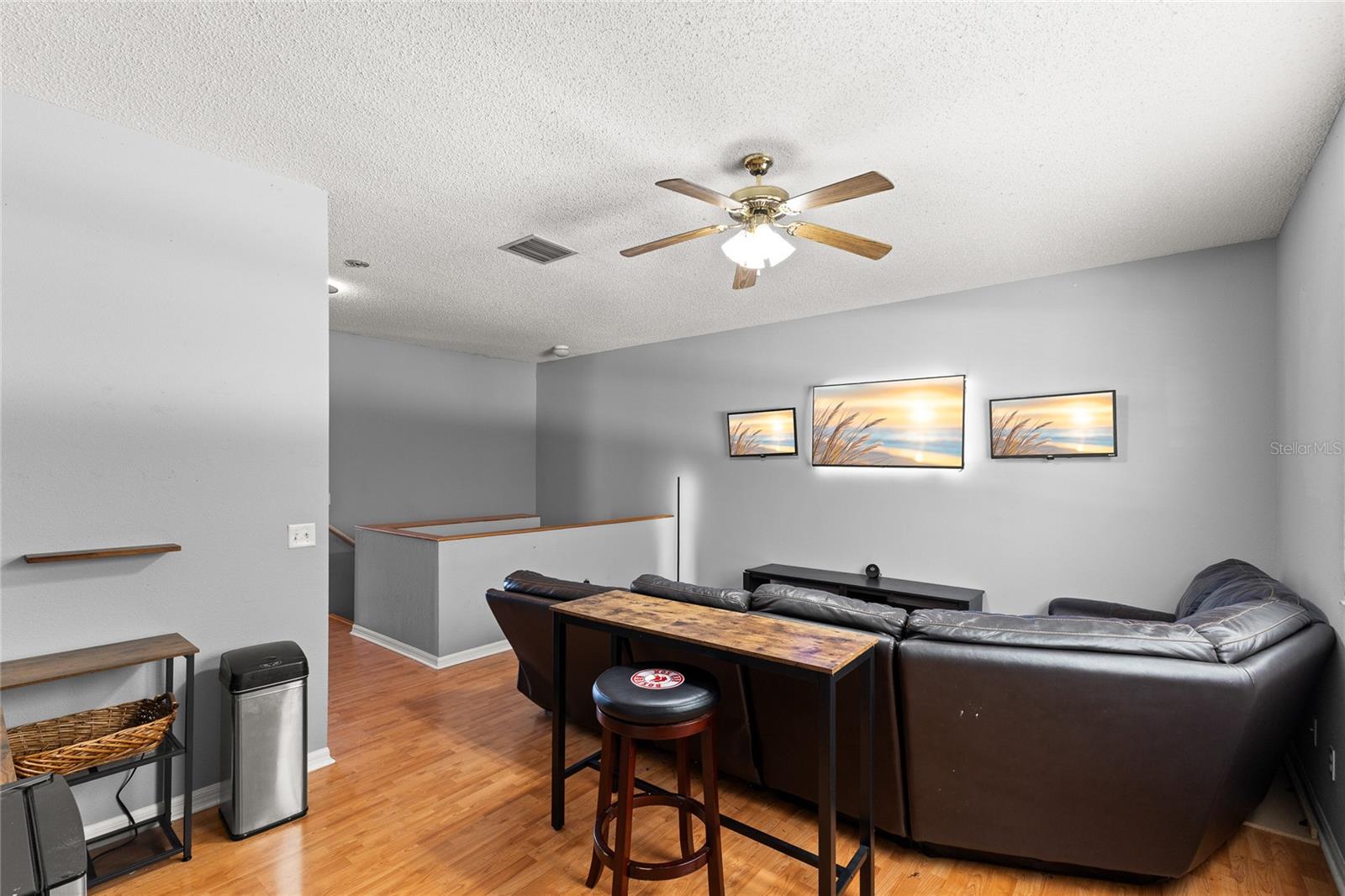 Second-floor loft/family room with warm wood-look flooring, neutral paint tones, recessed lighting, and a ceiling fan with light kit. The open layout overlooks the staircase below and offers flexible space for a media room, home office, playroom, or additional lounge area.