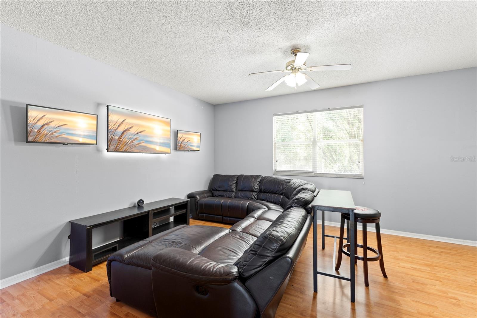 Second-floor loft/family room with warm wood-look flooring, neutral paint tones, ceiling fan with light kit, and oversized window bringing in natural light. Flexible layout offers space for a media room, gaming area, home office, or additional lounge seating.