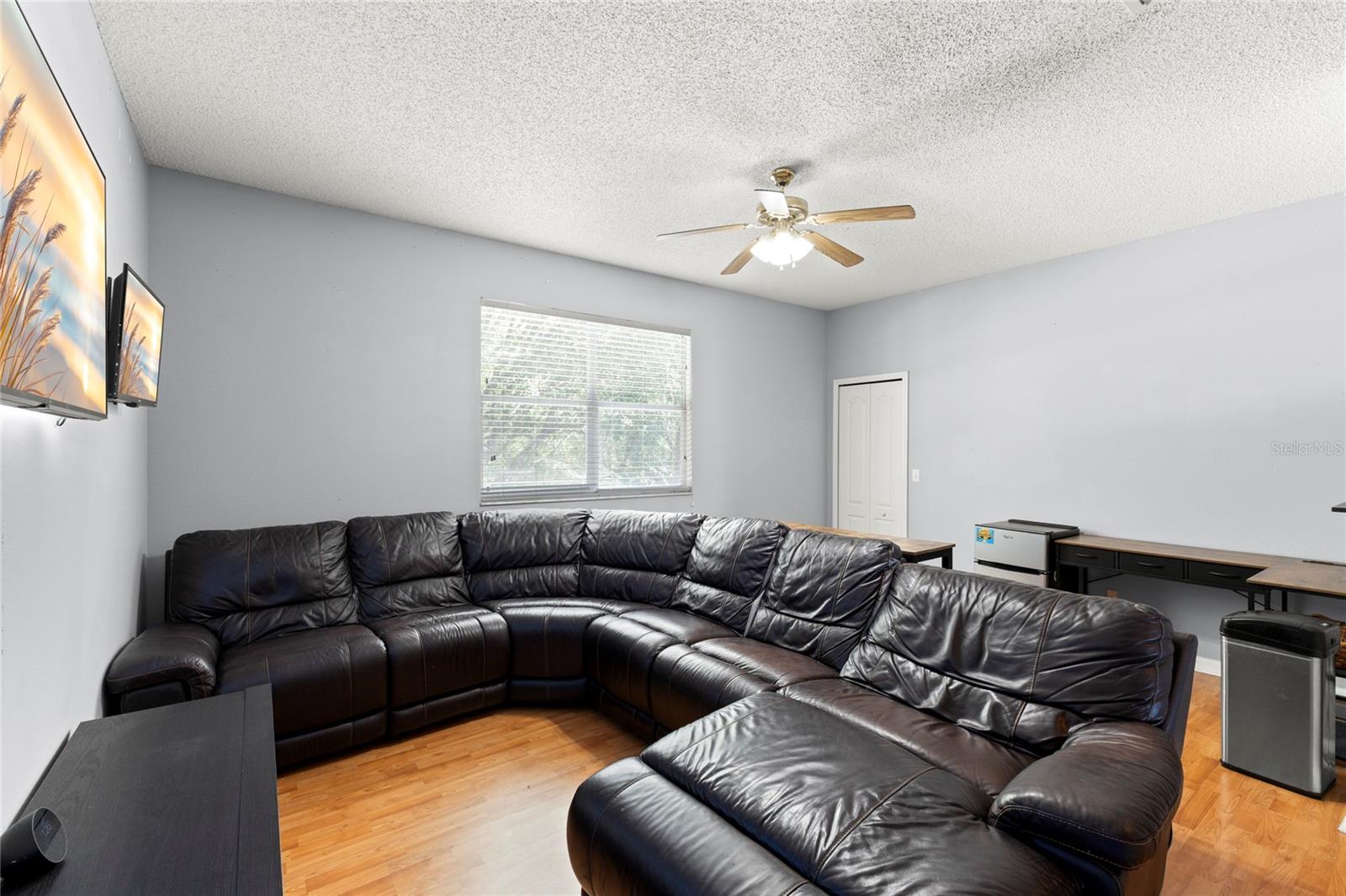 Second-floor loft/media room with warm wood-look flooring, neutral paint tones, ceiling fan with light kit, and oversized window with natural light. Flexible open layout accommodates sectional seating, media setup, gaming, or work-from-home space, with additional storage available in the walk-in closet.